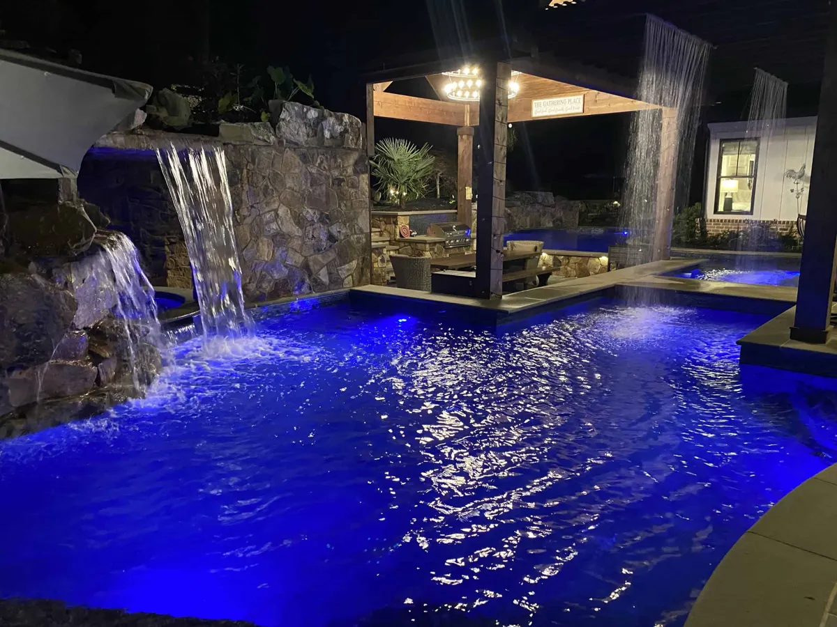 A lit backyard pool at night with stone waterfalls and a covered outdoor living area