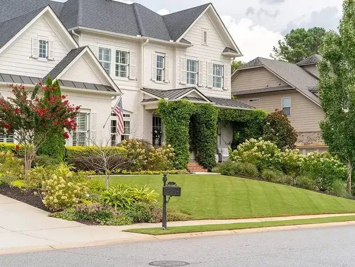 A white two-story home with a striped green lawn, flowering crepe myrtle and full planting beds