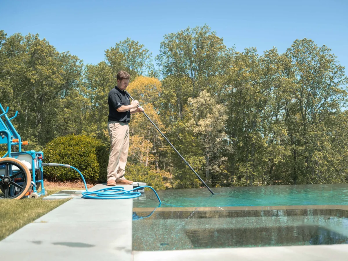 A Well Done Pools technician skimming an infinity-edge pool with trees behind it